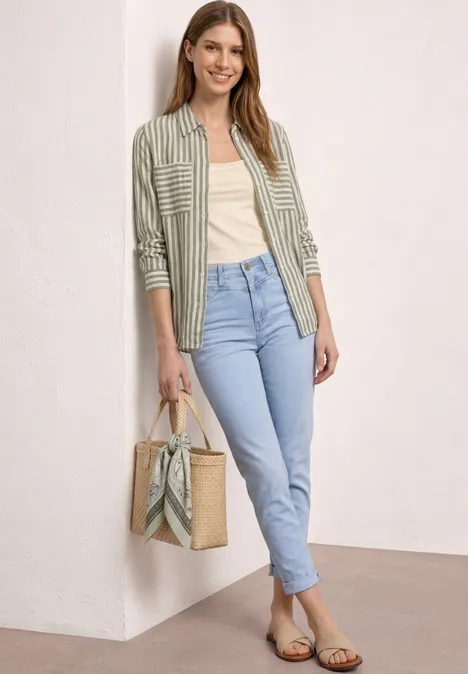 Frau in grün gestreiftem Hemd, Top, hellblauen Jeans, Sandalen und Strohtasche vor weißer Wand.