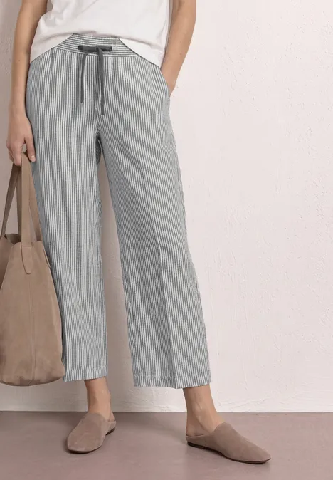 Frau trägt graue, gestreifte, weite Leinenhose mit Tunnelzug, weiße Bluse, beige Slipper und Tasche.