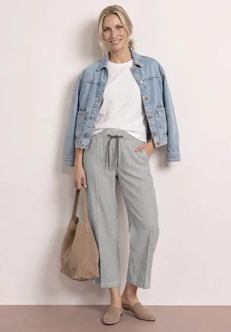 Frau trägt weißes T-Shirt, hellblaue Jeansjacke, graue weite Hose, beigen Wildleder-Beutel und Slip-Ons.