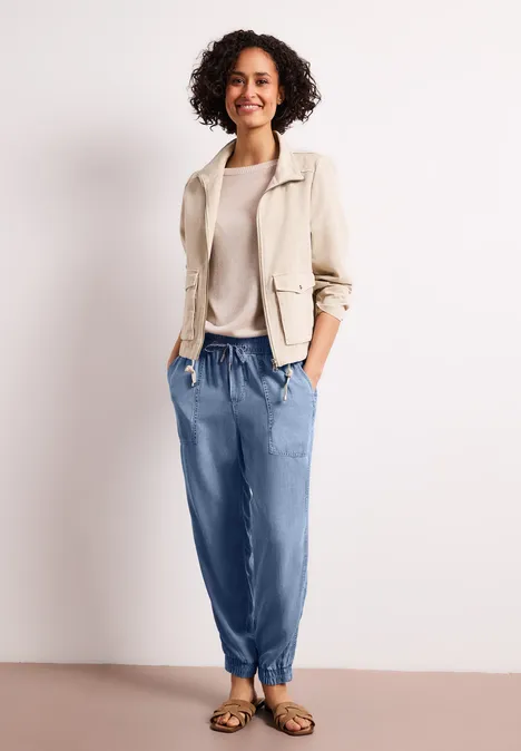 Vrouw in beige blazer, lichte trui, blauwe joggingsbroek en sandalen, handen in de zakken.