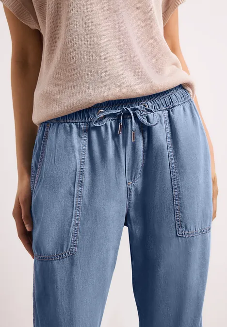 Persoon draagt losse, lichtblauwe jeans met taillekoord en grote zakken, gecombineerd met een beige trui.