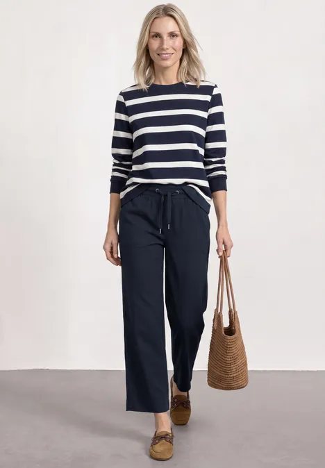 Straight Leg Hose mit Turn-Up-Detail deep blue Frau mit blau-weiß gestreiftem Shirt, weiten dunklen Hosen, Basttasche und Loafers.