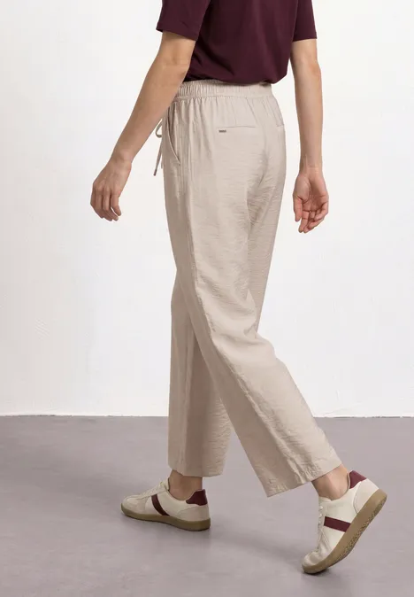Person im Gehen, trägt beige Leinenhose, cremefarbene Slipper mit braunem Riemen und weinrotes Oberteil.