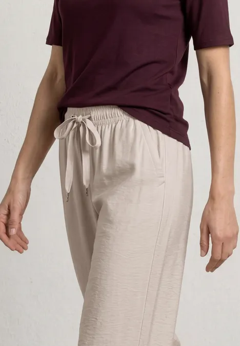 Person trägt helle Leinenhose mit Tunnelzug und dunkelrotes Shirt, vor schlichtem, hellem Hintergrund.