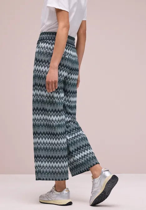 Een persoon draagt wijde, patroonbroek, een wit T-shirt en zilverkleurige sneakers tegen een lichte achtergrond.