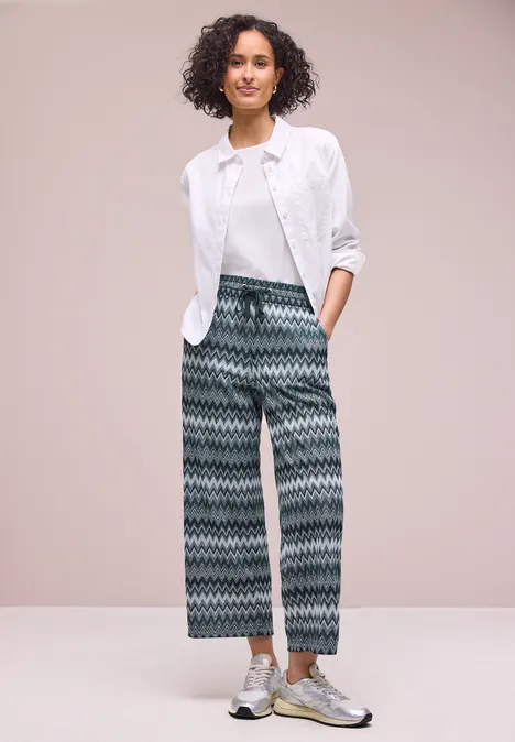 Persoon voor lichte achtergrond, witte blouse op t-shirt, wijde broek met zigzagmotief en metallic sneakers.