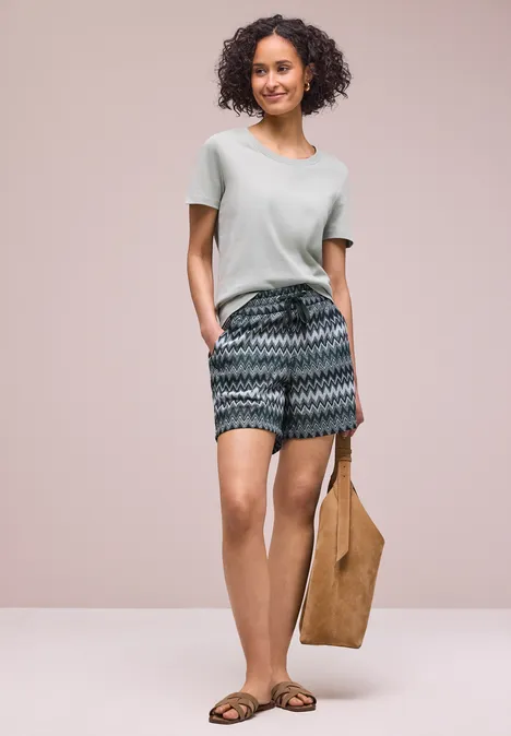 Frau in leichtem T-Shirt, gemusterten Shorts, flachen Sandalen und brauner Tragetasche vor schlichtem Hintergrund.