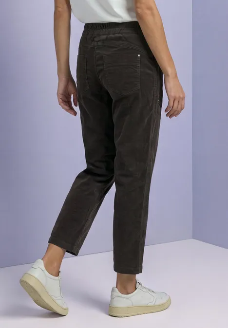Slim Leg Cordhose im Loose Fit black coffee Eine Person trägt schwarze Cordhose und weiße Sneakers vor lila Hintergrund, Rückansicht.