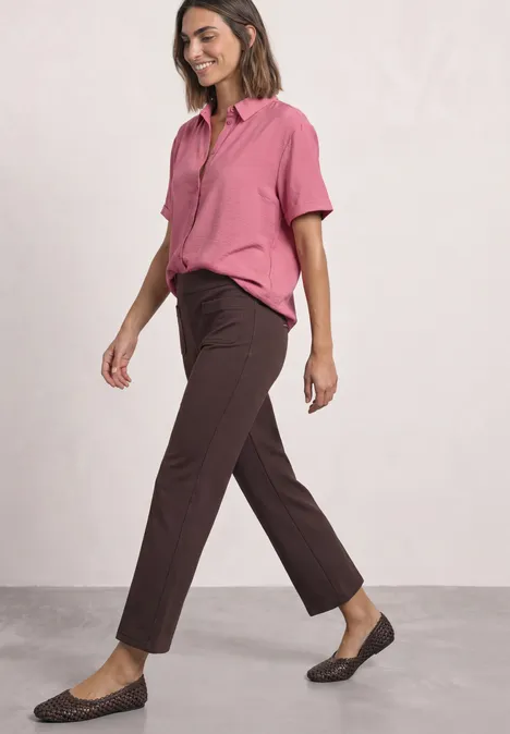 High Waist Straight Leg Hose im Casual Fit ruby vine Frau in rosa Hemd, dunkelbrauner Hose und flachen Schuhen geht seitlich vor neutralem Hintergrund.