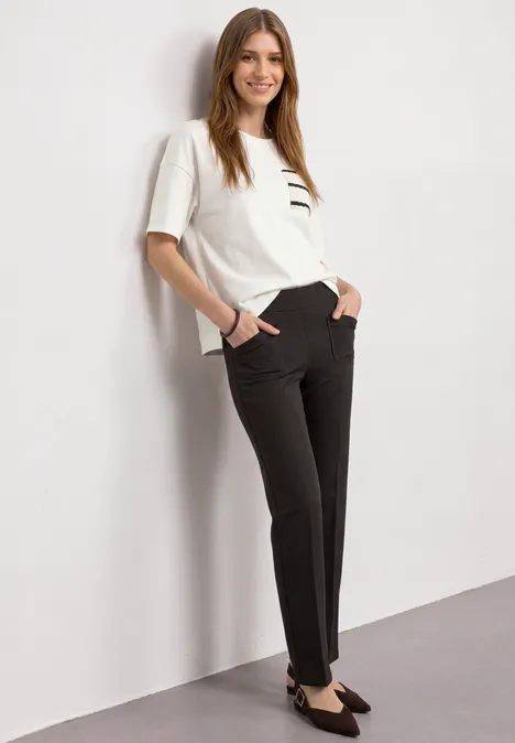 High Waist Straight Leg Hose im Casual Fit black coffee Frau lehnt an Wand, trägt weiße Bluse, schwarze Hose und schwarze Schuhe.