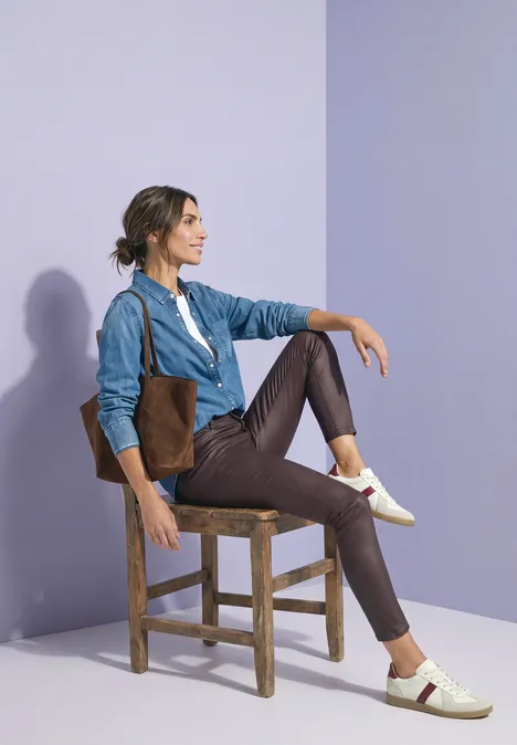 Pantalon taille moyenne, coupe slim, aspect cuir ruby vine Eine Frau sitzt auf einem Holzstuhl, trägt Jeansjacke, braune Hose und weiße Sneakers vor violettem Hintergrund.