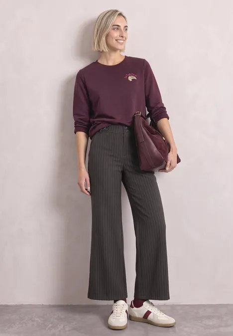 Frau in legerem Chic: burgunderfarbenes Shirt, gestreifte schwarze Schlaghose, weiße Sneaker, bordeauxfarbene Tasche.