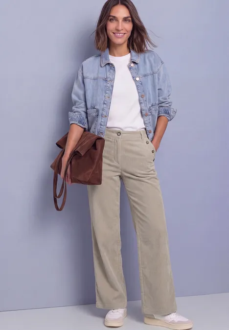 Eine Frau trägt Jeansjacke, weißes Shirt, weite beige Hose und hält eine braune Tasche.