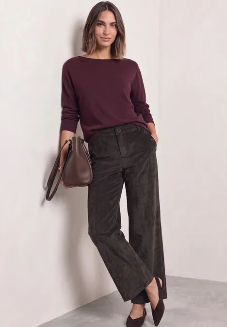 Frau lehnt an Wand, trägt bordeauxfarbenen Pullover, weite dunkelbraune Hosen und hält eine Tasche.