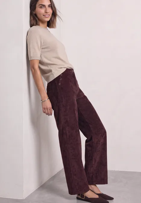 Frau lehnt an Wand, trägt beige T-Shirt und bordeauxrote, weit geschnittene Cordhose.