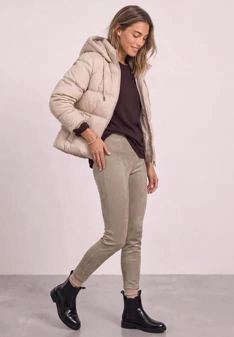 Frau in beiger Steppjacke, dunklem Pullover, hellen Hosen und schwarzen Stiefeletten, stehend vor hellem Hintergrund.