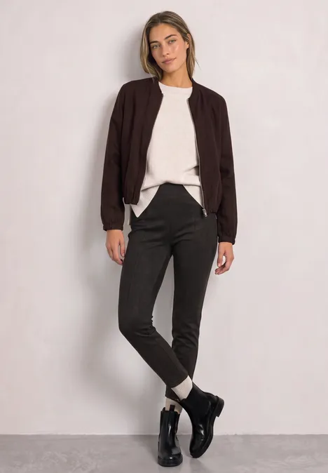 Eine junge Frau steht lässig vor hellem Hintergrund, trägt schwarze Jacke, weißen Pullover, schwarze Hose und Boots.