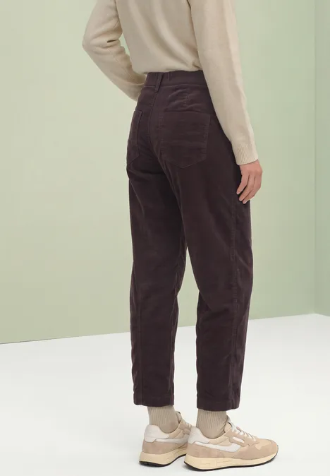 Person von hinten in violetter Cordhose, beigem Pullover und hellen Sneakers vor schlichtem Hintergrund.