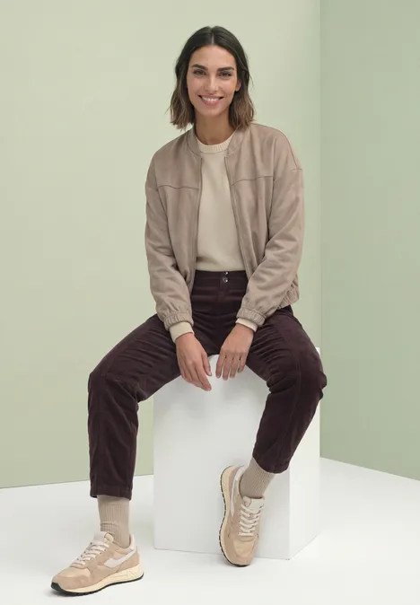 Frau in beige Jacke, weißem Shirt, braunen Cordhosen und beigen Sneakern sitzt auf weißem Würfel vor grünem Hintergrund.