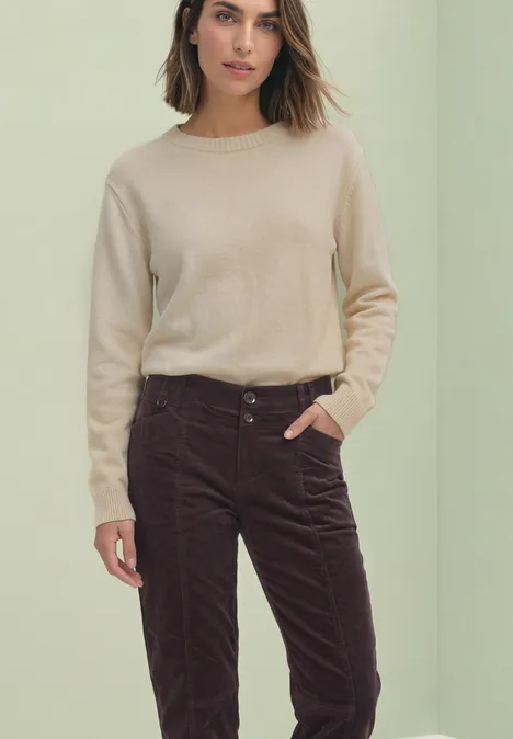 Eine Frau trägt einen beigen Pullover und dunkelbraune Cordhose, Hand in Tasche.