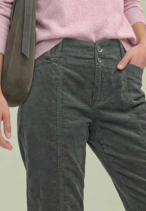 Frau trägt graugrüne Cordhose, rosa Pullover und grüne Umhängetasche (Ansicht bis Hüfte).