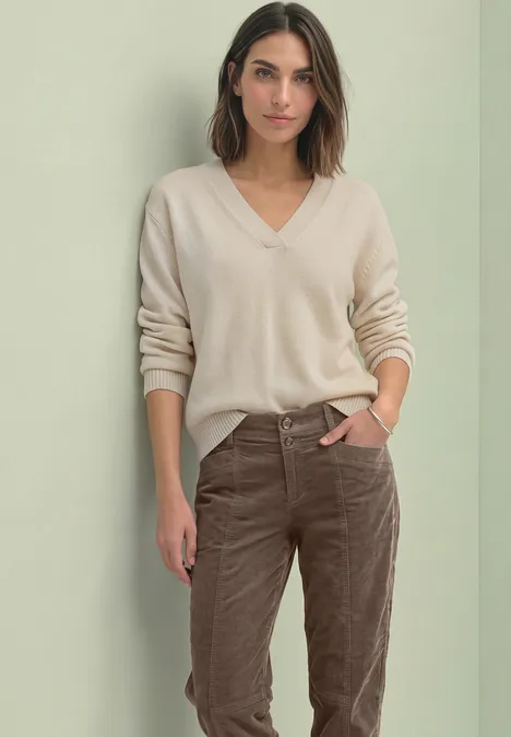Barrel Leg-broek van corduroy in casual pasvorm velvet mocha Vrouw in beige V-trui en bruine corduroy broek, hand in zak, staand voor een neutrale achtergrond.