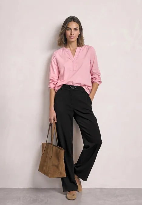 Vrouw in roze blouse en zwarte wijde broek met een bruine tas, staand voor een lichtgrijze achtergrond.