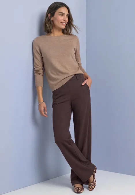 High Waist Straight Leg Hose im Casual Fit ruby vine Frau lehnt an lila Wand, trägt beigem Pullover, braune Weitehosen und Leo-Mokassins.
