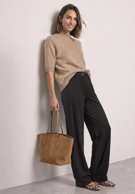 Vrouw in beige trui, zwarte wijde broek, suède tas en luipaardprint ballerina’s leunt tegen een muur.