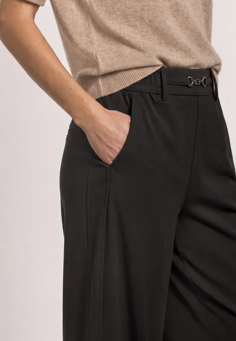 Zwarte, getailleerde broek met vouwen, beige top, hand in de zijzak.