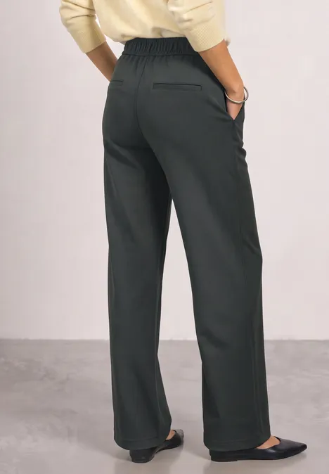 Vrouw van achteren in wijde zwarte high-waist broek, zwarte platte schoenen en crèmekleurige top.
