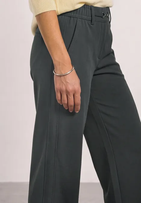 Persoon draagt donkergrijze broek en crème top, hand met zilveren armband zichtbaar.