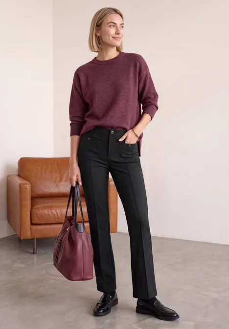 Bootcut Hose mit aufgesetzten Taschen Black Frau in burgunderrotem Pullover, schwarzer Hose und Schuhen, mit passender Tasche, steht neben braunem Sessel.