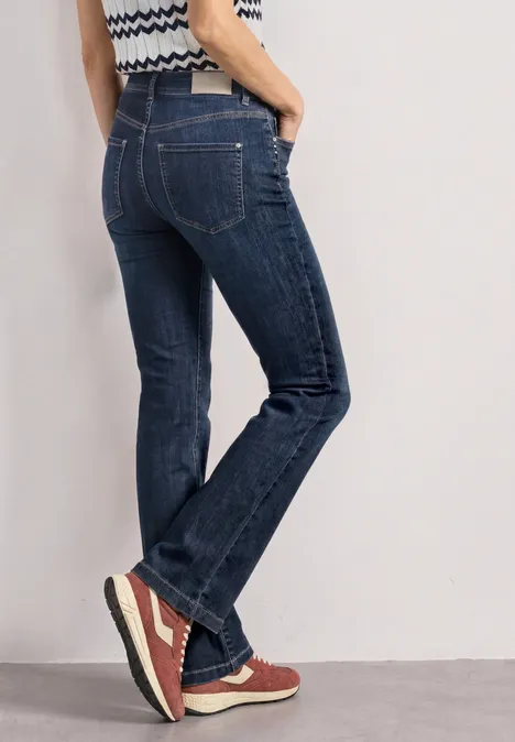High Waist Bootcut Jeans im Slim Fit Dark Blue Washed Person im blauen Jeanshosen und roten Sneakern, Seitenansicht, steht vor hellem Hintergrund.