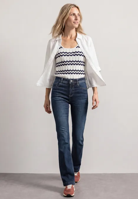 High Waist Bootcut Jeans im Slim Fit Dark Blue Washed Frau mit offenem weißen Hemd über gestreiftem Top, dunklen Jeans und Sandalen.
