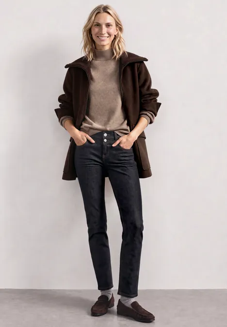 Dżinsy z Mid Waist i wąskimi nogawkami o Casual Fit Deep Rinse Washed Kobieta w brązowym płaszczu, swetrze z golfem, czarnych dżinsach i mokasynach na prostym tle.