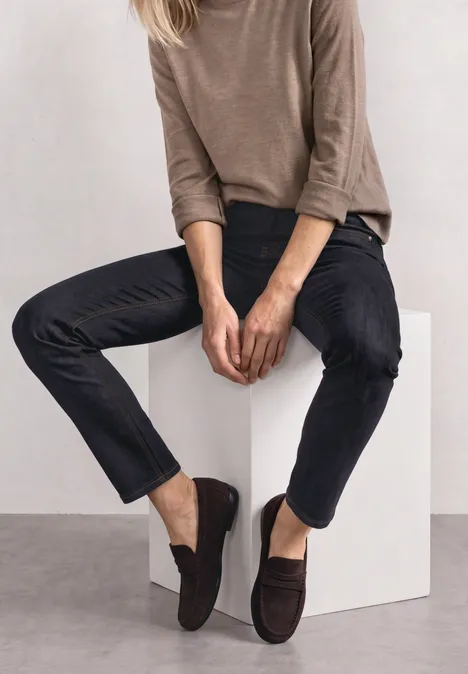Dżinsy z Mid Waist i wąskimi nogawkami o Casual Fit Deep Rinse Washed Osoba siedzi na białym sześcianie, nosi jasnobrązowy sweter, ciemne dżinsy i brązowe mokasyny.