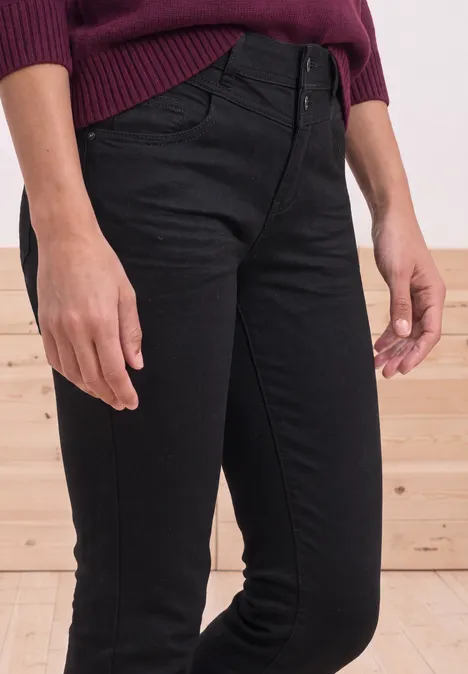 Zwarte high-waist skinny jeans met knoopsluiting, gecombineerd met een bordeauxrode trui voor een houten wand.