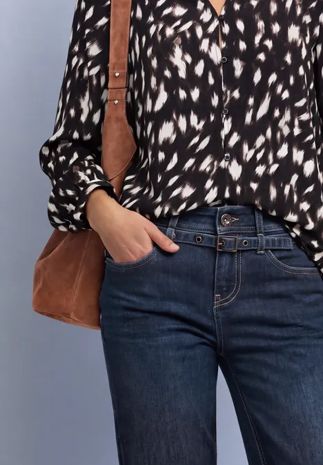 Frau mit schwarz-weiß gemusterter Bluse, blauen Jeans und Umhängetasche, eine Hand in der Tasche.