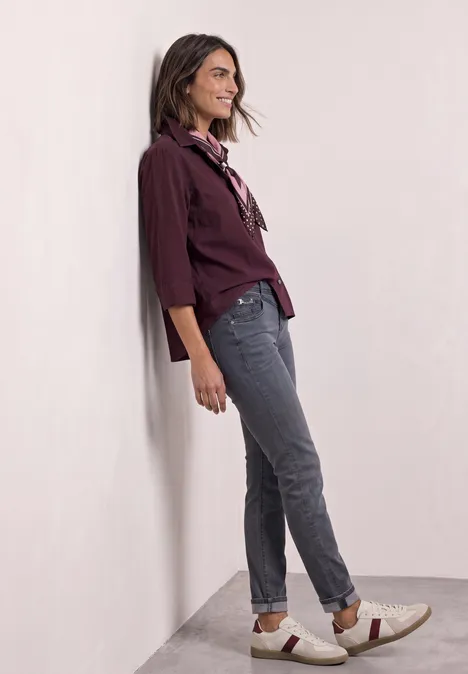 Frau lehnt lächelnd an Wand, trägt weinrote Bluse, graue Jeans und bequeme Slipper.