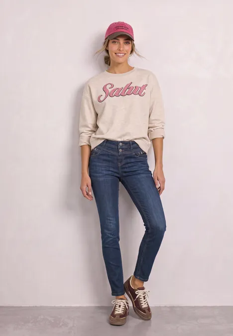 Frau in Jeans, beigem Sweatshirt mit 'Salut'-Aufdruck, rosa Cap und braunen Sneakern vor neutralem Hintergrund.