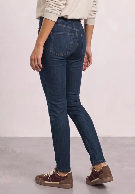 Person trägt dunkle Skinny-Jeans und braune Schuhe, steht seitlich vor heller Wand.