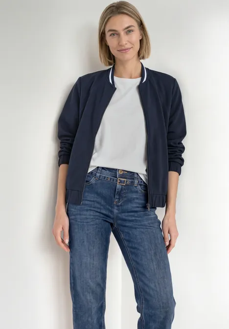 Straight Leg Jeans mit Gürtel Mid Blue Random Washed Eine lächelnde Frau im navyblauen Blouson, weißem T-Shirt und Jeans steht vor hellem Hintergrund.