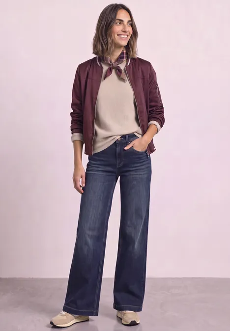 High Waist Wide Leg Jeans im Loose Fit Dark Blue Clean washed Frau in Burgunder-Bomberjacke, beigefarbenem Pullover, weiten Jeans und beigen Schuhen vor hellem Hintergrund.