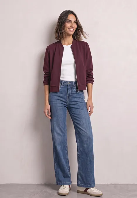 High Waist Wide Leg Jeans im Loose Fit Medium Blue Soft Washed Frau trägt burgunderfarbenen Blouson, weißes Shirt, blaue Jeans und weiße Sneaker vor hellem Hintergrund