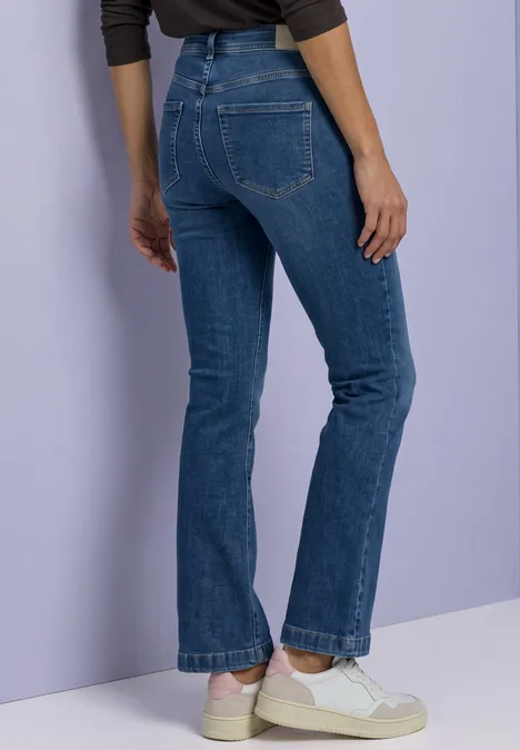 Eine Person steht seitlich, trägt blaue Jeans und weiße Sneaker vor einem hellvioletten Hintergrund.