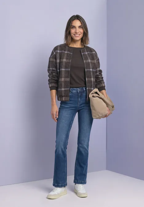Eine lächelnde Frau trägt karierte Jacke, Jeans und weiße Sneaker, hält eine beige Tasche vor lilafarbenem Hintergrund.