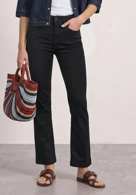 Person trägt schwarze Jeans, Jeansjacke, weiße Bluse, braune Sandalen und hält eine mehrfarbige gestreifte Tasche.