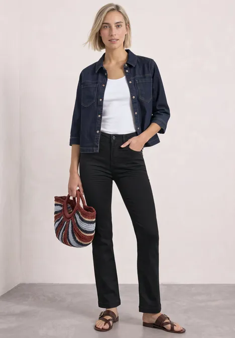 Frau in Jeansjacke, weißem Top, schwarzen Hosen und Sandalen hält bunte Tasche.