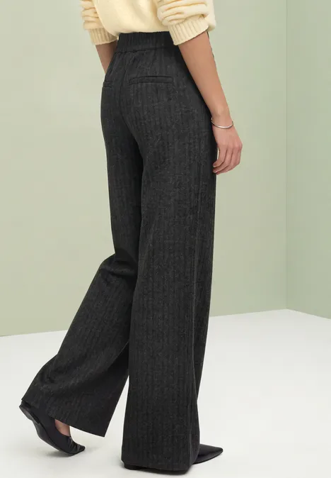 Achteraanzicht van persoon in donkergrijze pantalon met krijtstrepen, wijde pijpen, gecombineerd met zwarte hakken.